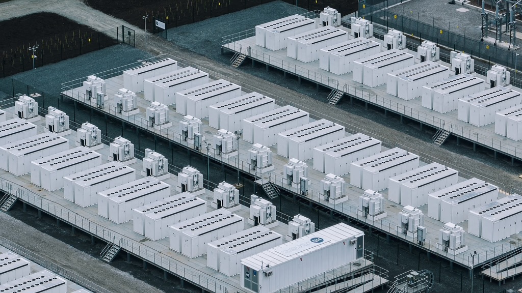 Aerial view of a grid-scale battery storage site: long rows of white
                  container-sized battery units on raised platforms, linked by access
                  walkways and stairs, with a substation at the top of the frame.